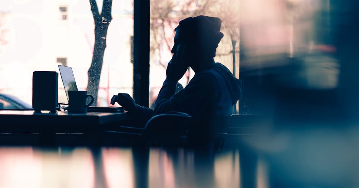 A silhouetted figure working on a laptop at a table by a window